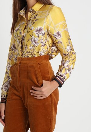 Femme portant un chemisier jaune à manches longues à motif floral, rentré dans un pantalon en velours côtelé rouille taille haute, la main dans la poche.