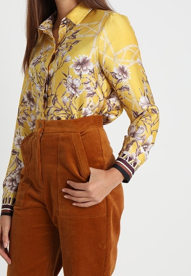 Femme portant un chemisier jaune à manches longues à motif floral, rentré dans un pantalon en velours côtelé rouille taille haute, la main dans la poche.