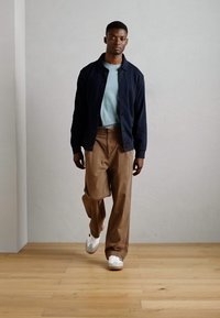 Cardigán navy, camiseta azul claro, pantalones marrones y zapatillas blancas. El modelo está de pie sobre un suelo de madera frente a una pared lisa.