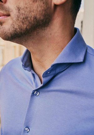 Hals en kaaklijn van een man met een korte baard, die een lichtblauw overhemd met open kraag draagt.