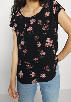 Femme portant un haut noir à manches courtes avec des motifs floraux roses et beiges, associé à un jean bleu clair, debout avec une main sur la hanche.