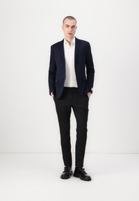 Marineblazer over een wit shirt, gecombineerd met zwarte broek en zwarte schoenen. Slim fit, gladde stof. Handen in de zakken, staande pose.