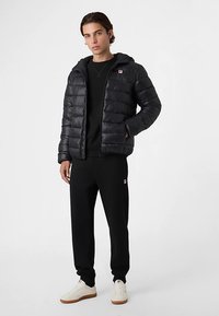 Giacca imbottita nera con cappuccio, con pannelli trapuntati; indossata sopra una maglietta nera e abbinata a pantaloni della tuta neri e sneakers bianche.