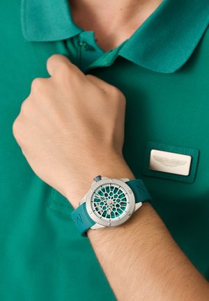 Srebrna ura z zeleni silikonski pasom ima geometrijski izrez na obrazu in je oblečena čez zeleni polo majico.