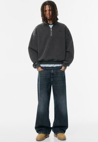 Przesadny czarny sweatshirt z zamkiem błyskawicznym, nałożony na niebieską koszulę, połączony z szerokimi ciemnoniebieskimi dżinsami i brązowymi butami.