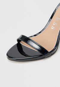 Sandalia de tacón alto de charol negro con punta puntiaguda, con una sola tira ancha sobre el arco y una plantilla beige.