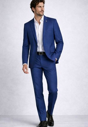 Homme en costume bleu ajusté, chemise blanche, ceinture noire et chaussures de ville noires, posant avec une main dans la poche devant un fond uni.