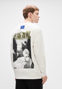 Λευκό βαμβακερό μπλουζάκι με μακριά μανίκια που έχει μια ασπρόμαυρη εκτύπωση γραφικών στο πίσω μέρος, παρουσιάζοντας χαρακτήρες και στυλιζαρισμένο κείμενο.