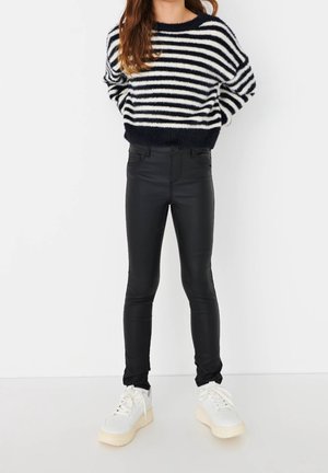 Donna che indossa pantaloni neri aderenti, sneakers bianche e un maglione fluffy a righe bianche e nere, in piedi davanti a uno sfondo bianco uniforme.