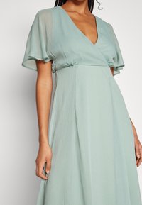 Robe plissée vert clair avec décolleté en V et manches frivoles. Tissu doux et fluide avec une texture subtile et une silhouette gracieuse.