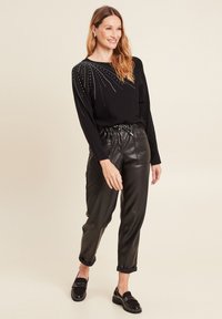 Schwarzer Strickpullover mit silbernem Nietenmuster auf einer Schulter, kombiniert mit schwarzen Kunstlederhosen und schwarzen Loafers.