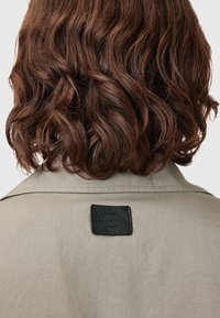 Beige trenchcoat med tekstureret stof, der har et rektangulært sort lædermærke på ryggen med prægede bogstaver.