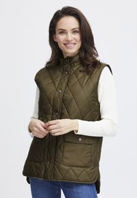 Grüne gesteppte Weste mit hohem Kragen, Druckknöpfen und seitlichen Taschen, getragen über einem weißen Langarmshirt, kombiniert mit blauen Jeans.