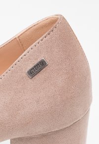Gros plan sur une chaussure à talon bloc en daim beige avec une plaque métallique de la marque "mtng" près du col, montrant le matériau texturé et les détails de la couture.