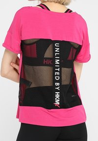 Osoba oblečená v jasně růžovém tričku s černou síťovinovou panely na zádech se nápisem "UNLIMITED BY HKMX," zkombinovaném s černými legínami.