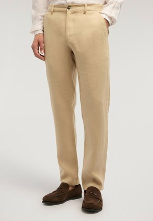 Beige bukser med lige pasform kombineret med brune mokkasiner i ruskind, med den ene hånd i lommen, båret med en lys, stribet skjorte.