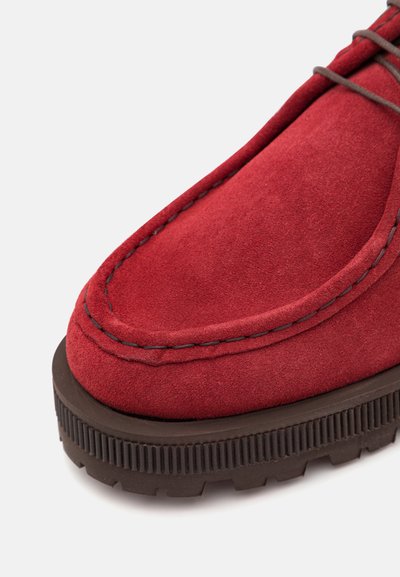Crvena suede cipela s kontrastnim tamnosmešnim gumenim đonom, zaobljenim prednjim delom, detaljno prošivena duž ivice i teksturiranom površinom.