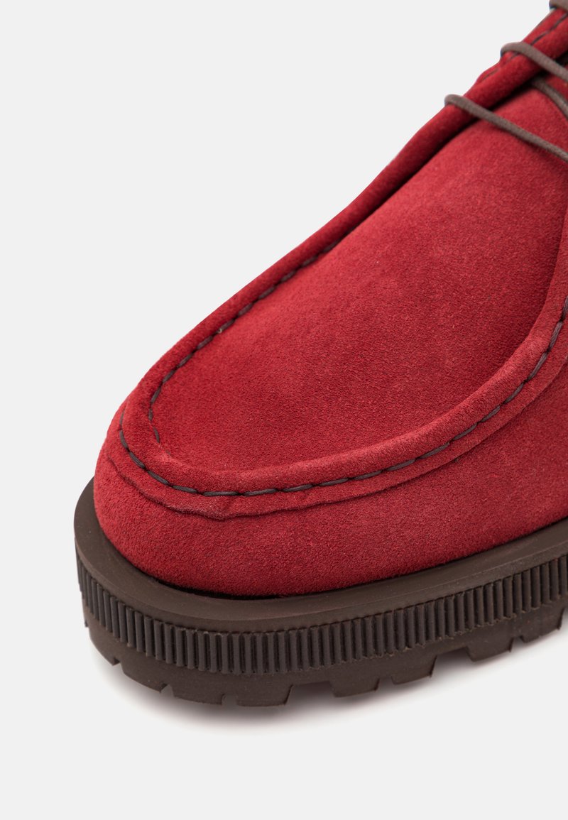 Scarpa in suede rosso con suola in gomma marrone scuro a contrasto, punta arrotondata, cuciture dettagliate lungo il bordo e superficie testurizzata.