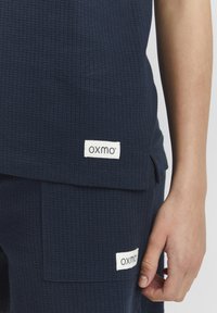 Marineblå, struktureret top med en rektangulær lomme og et hvidt mærke med teksten "oxmo." Materialet ser let og åndbart ud.