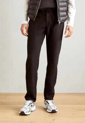 Zwarte linnen broek met een rechte pasvorm, trekkoord in de taille en subtiele textuur, gecombineerd met grijze sportschoenen en een gewatteerde vest.