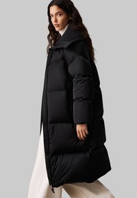 Abrigo de plumas negro con cuello alto, que presenta secciones acolchadas, mangas largas y un cierre frontal con cremallera. Textura suave y corte oversized.