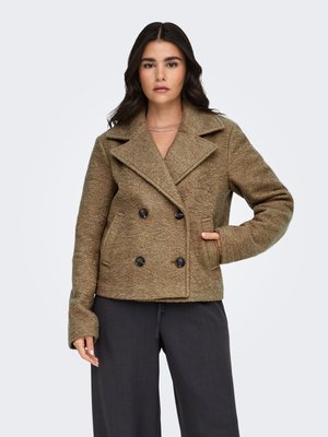 Femme portant un manteau marron texturé à double boutonnage avec des boutons noirs et un pantalon noir à jambes larges, debout avec une main dans la poche.