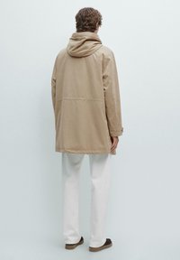 Beige huvad kappa med en avslappnad passform, som har en enkel design, texturerad tyg och en längd till mitten av låret, i kombination med vita byxor.