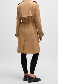 Osoba ubrana w beżowy trencz do kolan z paskiem, czarne legginsy i czarne skórzane mokasyny na obcasie, stojąca na jednolitym tle.