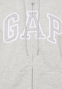 Felpa grigia con zip con logo "GAP" bianco con dettagli ricamati, cappuccio con coulisse e due tasche frontali. Morbido tessuto in misto cotone.