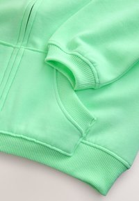 Manche et poche de sweat-shirt vert menthe avec poignets et ourlet côtelés, présentées en gros plan sur un fond neutre.