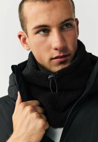 Čierny fleece nákrčník s nastaviteľným elastickým pásikom; v kombinácii s tmavým kabátom. Detailný záber textúry a vrstvených tkanín.