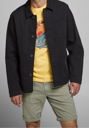 Mand iført en sort jakke med knapper over en gul T-shirt med grafik, kombineret med lysegrønne shorts med opkrogede ben mod en grå baggrund.