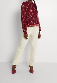 Blouse transparente à fleurs bordeaux avec manches longues et col fendu, associée à un pantalon crème à jambes droites et des escarpins rouges à bouts pointus.
