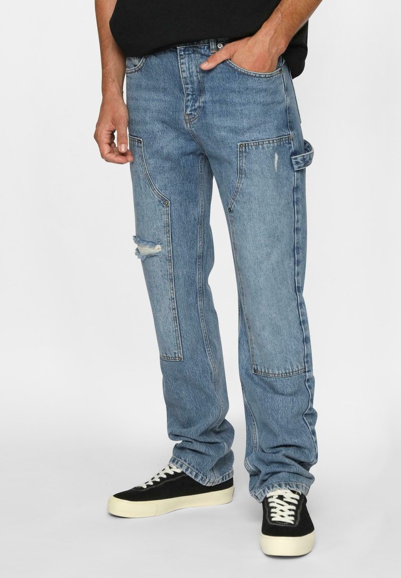 Lyseblå denimjeans med en avslappet passform, med flere lommer, et slitt kne og subtile sømdetaljer. Brukt med svarte joggesko.