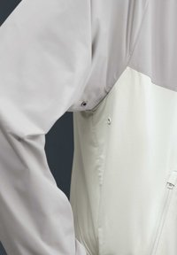 Primer plano del costado de una chaqueta que muestra una manga gris claro, cuerpo blanco hueso, ventilaciones con botones a presión bajo el brazo y un bolsillo vertical con cremallera.