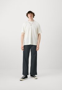T-shirt blanc à manches courtes, pantalon noir à coupe décontractée et basket hautes noires. Le modèle porte une casquette blanche et des colliers superposés.