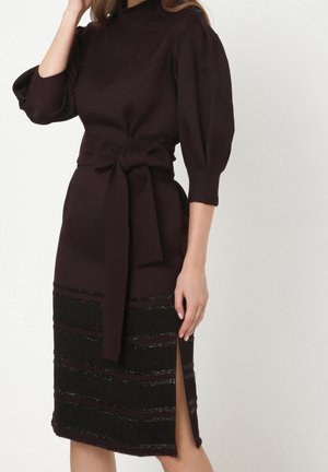 Nainen, jolla on päällään tummanvioletti neulemekko, jossa on pöyhistetyt hihat, suuri solmittu vyö vyötäröllä ja etuosassa teksturoidut vaakasuorat raidat.