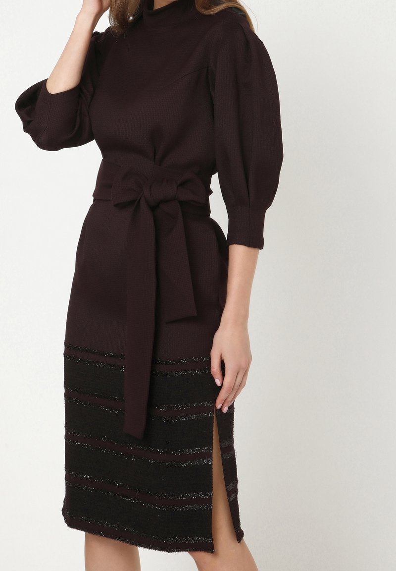 Femme portant une robe tricotée violette foncée avec des manches bouffantes, une large ceinture nouée à la taille et une fente avant avec des rayures horizontales texturées.