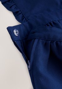 Detail tmavomodrého odevu s čiastočne otvoreným zipsom a priehľadným gombíkom na nariasenom okraji látky.