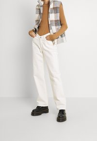 Gestrickte Weste in Grau, Weiß und Braun über einem gerippten braunen Pullover; kombiniert mit weißen Jeans und schwarzen Plateaustiefeln.