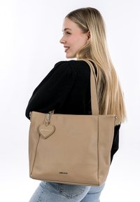 Beige nahkatoote kott tekstuuri viimistlusega, pikad sangad ja südamekujulise ripatsi. Omab sujuvat lukku ja minimaliseeritud brändingut.