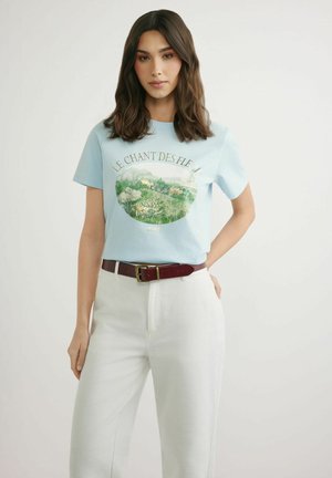 Kobieta z długimi ciemnymi włosami, ubrana w jasnoniebieski t-shirt z nadrukiem kwiatowego krajobrazu oraz białe spodnie z wysokim stanem i brązowym paskiem.