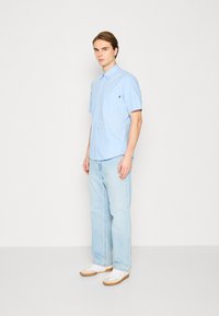 Camisa de botones de manga corta color azul claro con un bolsillo en el pecho, combinada con pantalones vaqueros azul claro y zapatillas blancas con suela en tono marrón.