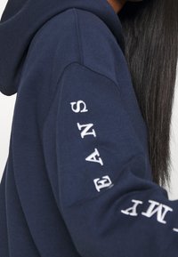 Mjukt texture i marinblå hoodie med vit text längs ärmen. Designad med en rymlig huva och ribbade ärmslut.
