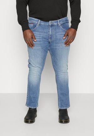 Mężczyzna w jasnoniebieskich, dopasowanych spodniach jeansowych, czarnej koszuli z długim rękawem i czarnych butach, stojący z rozluźnionymi rękami wzdłuż ciała.