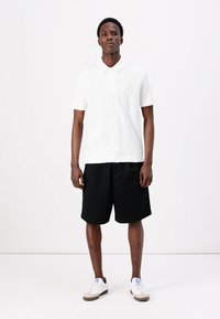 Balta īsām piedurknēm pogājamā krekla ar teksturētu rakstu kombinācija ar melnajām šortiem un baltām apaviem ar bežiem akcentiem. Attēlots ikdienišķs apģērbs.