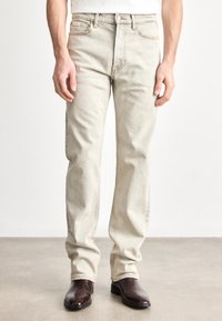 Gaiši bēšas džinsa bikses ar taisnu piegriezumu, ar priekšējām kabatām, pogas aizdari un redzamu šuvju izšuvumu. Kombinētas ar tumši brūnām zābakiem.