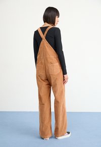 Brown corduroy overalls with wide legs and adjustable straps, featuring two back pockets. Worn over a black long-sleeve top and white sneakers.