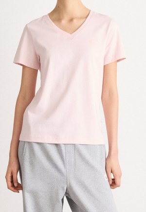 Persoon draagt een lichtroze V-hals T-shirt met korte mouwen en lichtgrijze casual broek tegen een effen witte achtergrond.