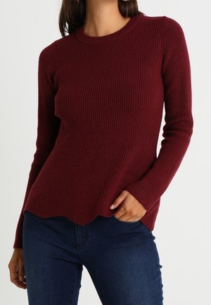 Vrouw draagt een nauwsluitende, geribbelde bordeauxrode trui met lange mouwen en een geschulpte zoom, gecombineerd met donkerblauwe jeans.